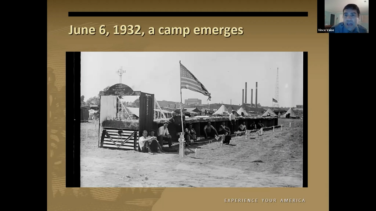 Black and white photo of men sitting in makeshift shelters with a U.S. flag waving out front.