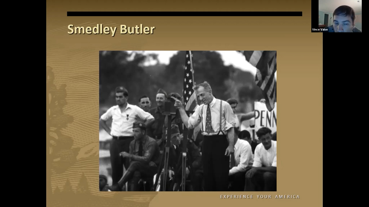 Black and white photo of a man in shirtsleeves up on a stage gesturing vigorously in front of a U.S. flag