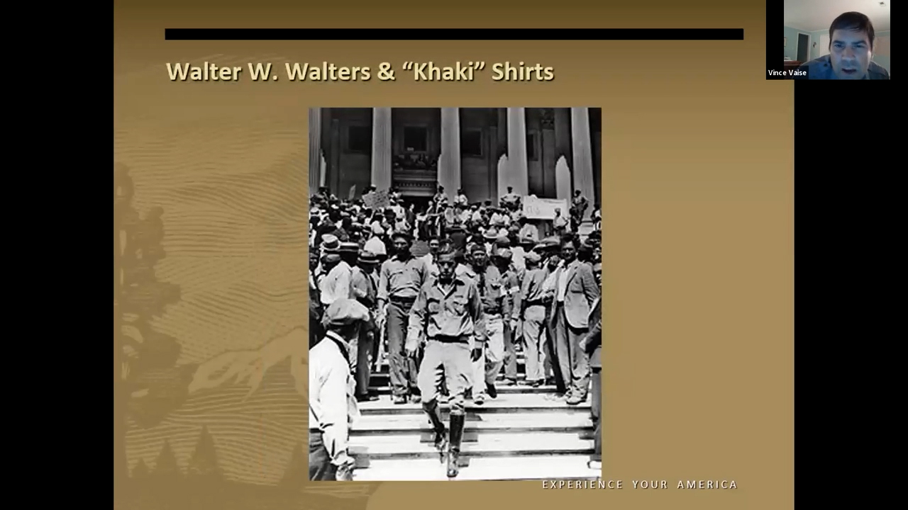 Black and white photo of a slender white man wearing tall boots, khaki pants and a khaki shirt descending the steps of a building followed by men dressed similarly
