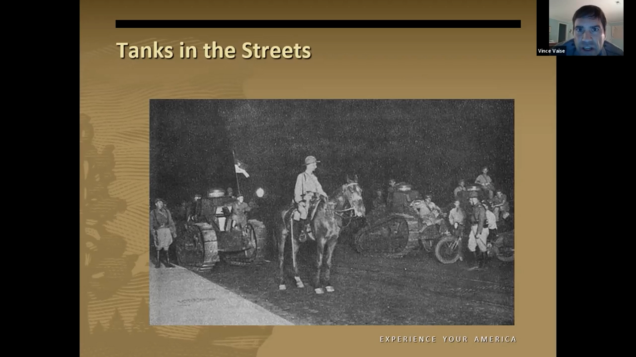 Black and white photo of soldiers on horseback amidst very small WWI tanks rolling down the street