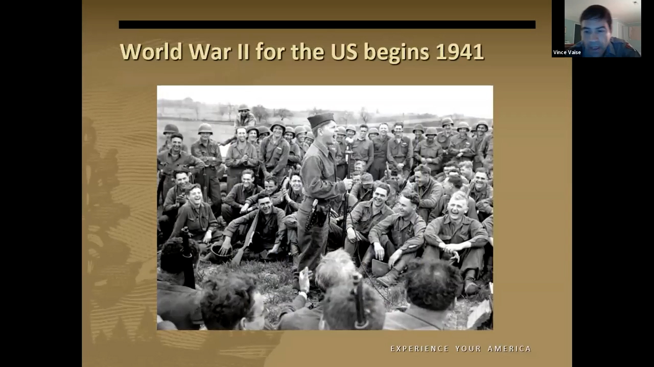 WWII soldiers sitting down in a grassy field listening to another soldier speaking into a microphone