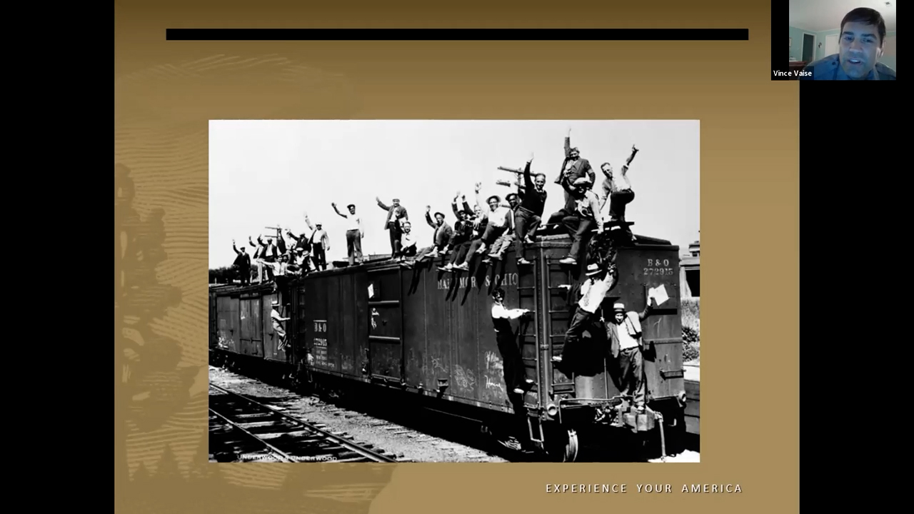 Black and white photograph of a train care covered with men standing on the roof and clinging to the sides