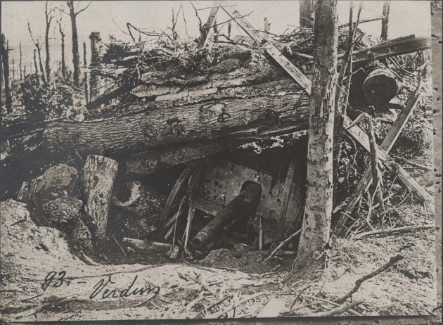 Black and white photograph of a destroyed German 77mm Howitzer under rubble, broken trees and dirt, at Verdun, France
