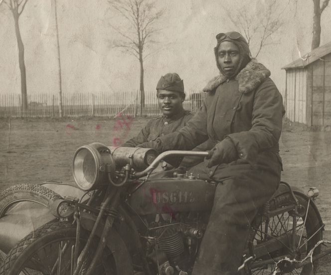 A black and white photo of an African American man dressed in a jumpsuit and gloves sits on a motorcycle, while a second man sits in the sidecar, wearing a uniform hat and wool jacket.
