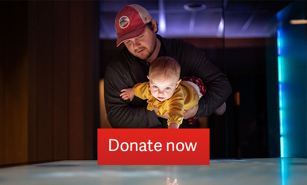 A white man holds a baby in his arms while the baby reaches out to touch the surface of an interactive table. Text: 'Donate now'