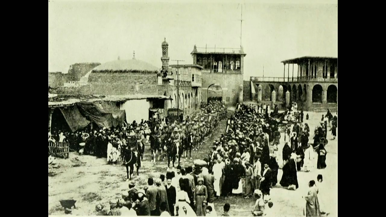 Black and white photograph of a column of horsed soldiers marching down a street while civilians look on
