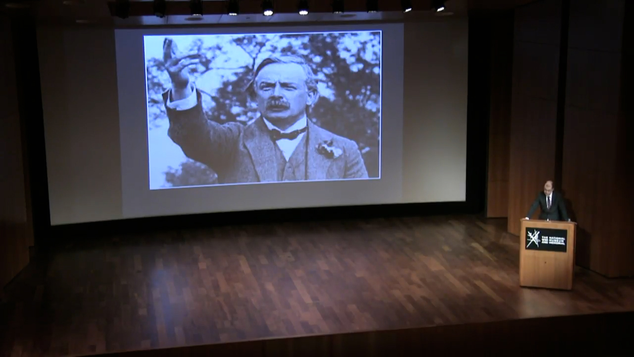 Black and white photo of a mustachioed white man in a civilian suit gesturing as he gives a speech