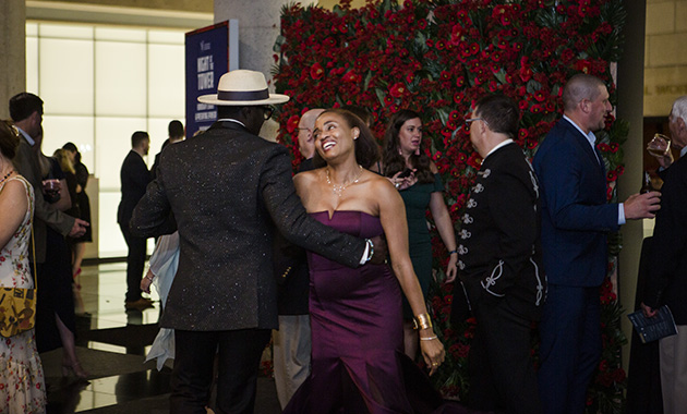 A Black woman in a cocktail dress dances with a Black man in a suit and white fedora, against a rose-covered photo backdrop