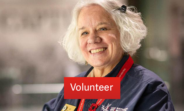 A white woman with short white hair wearing a blue volunteer shirt smiles at the camera. Text: 'Volunteer'