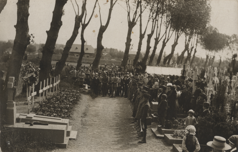 Black and white photo of Americans soldiers and French citizens standing in front of a row of white crosses. People are dressed formally and the white crosses are set with flowers.
