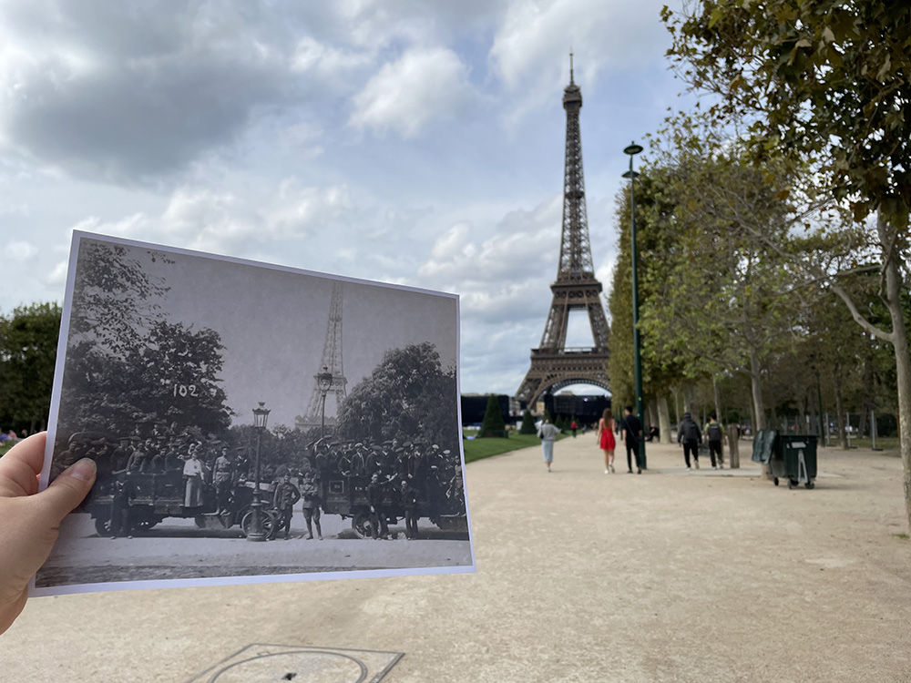 Modern photo of the Eiffel Tower. A person is holding up a black and white historical photo of WWI soldiers posing in front of the tower