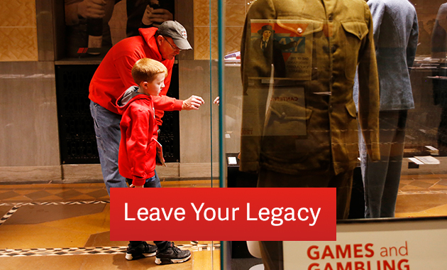 An adult in a red sweatshirt points something out in an exhibition case full of WWI uniforms to a child in a red sweatshirt. Text: 'Leave Your Legacy'