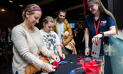 An adult woman and a younger girl craft poppy pins at a table while a Museum Volunteer looks on