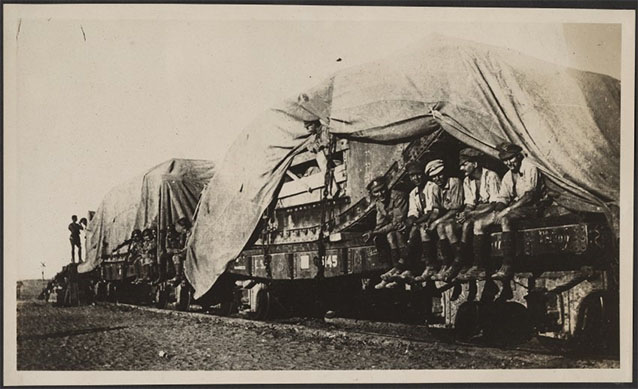 Black and white photo of two flatbed railroad cars loaded with tanks. A group of men are sitting on the edge of one of the cars.