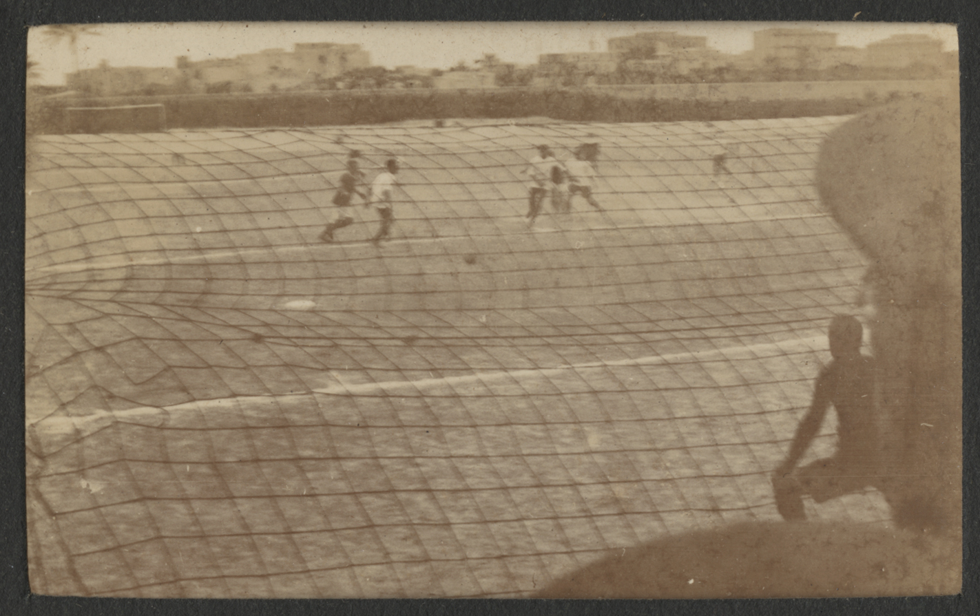Black and white photograph of people playing soccer in a field. The photo is taken through a net. There is part of the back of a person's head, wearing a hat, in the foreground.