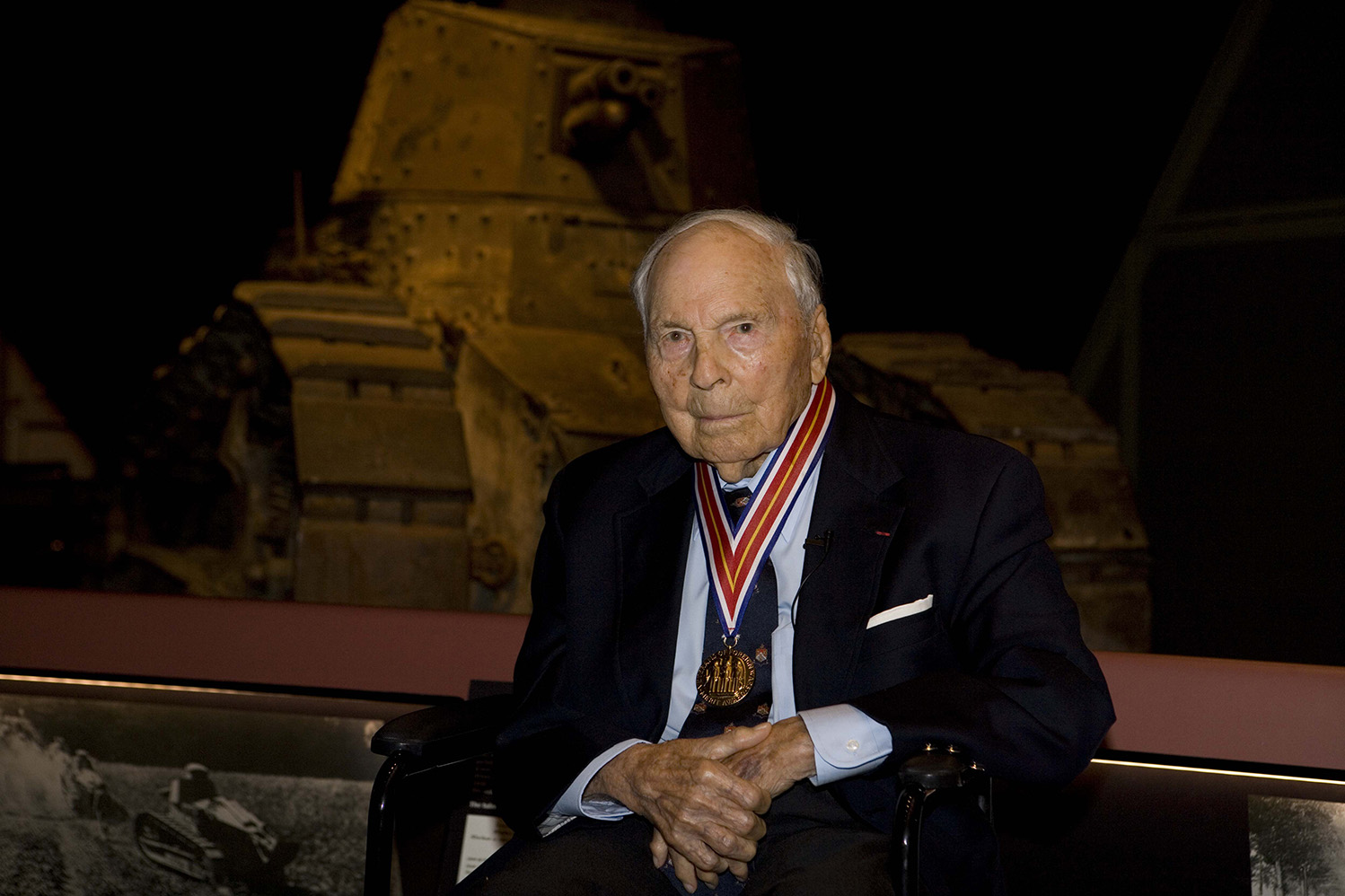 An elderly white man wearing a suit and a medal around his neck is seated in front of a museum exhibit of a WWI tank.