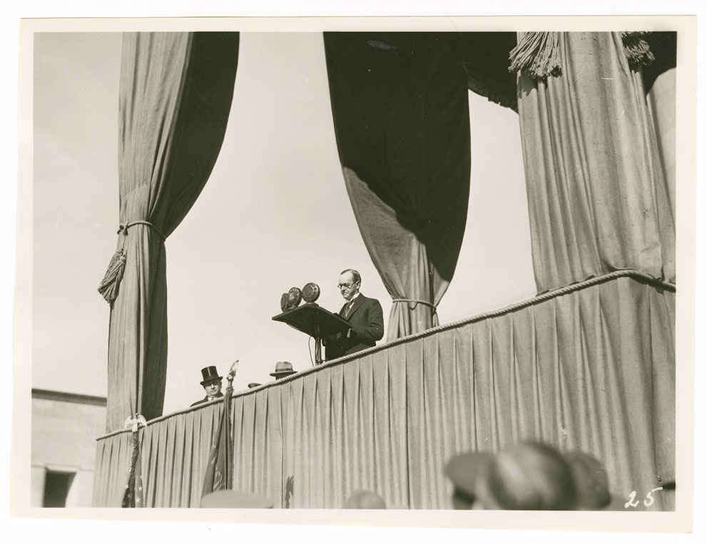Black and white photo of a white man in a suit standing at a podium on a large canopied platform.