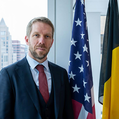 Middle-aged white man with close-cropped hair and beard wearing a dark suit, standing in front of the Belgian and U.S. flags