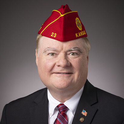 Older white man in a black suit wearing a red cap with yellow detailing