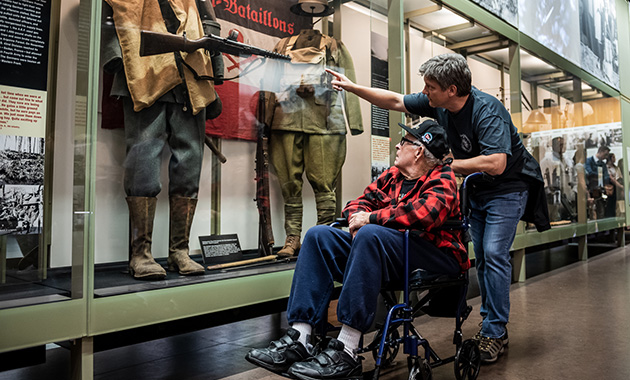 Modern photo of an older white man sitting in a wheelchair in a museum gallery. A younger white man points something out in an exhibition case while pushing the wheelchair.