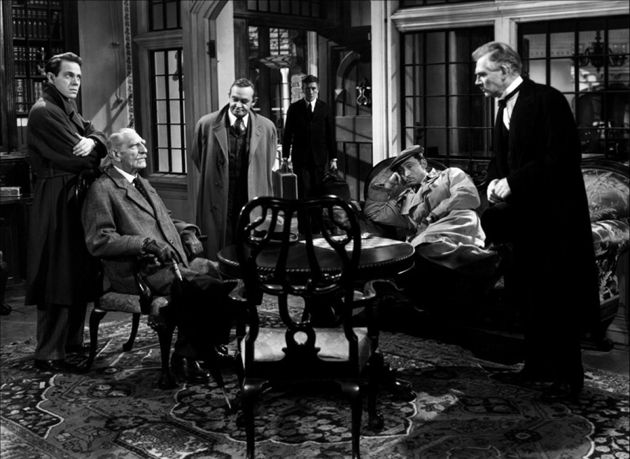 Black and white film still showing six men dressed in vintage style standing or seated in an elegant living room