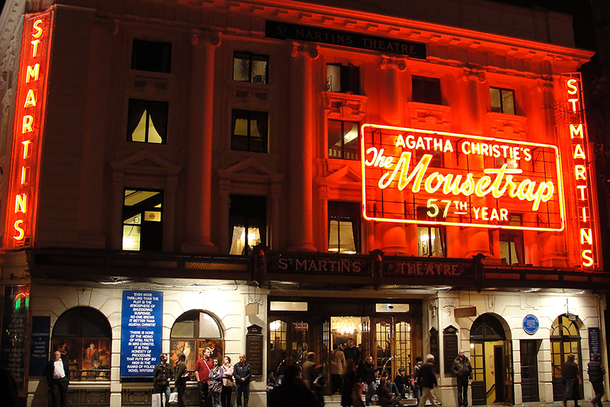 Color photo of the front of a theater lit up red at night. A large neon sign reads 'Agatha Christie's The Mousetrap 57th Year'
