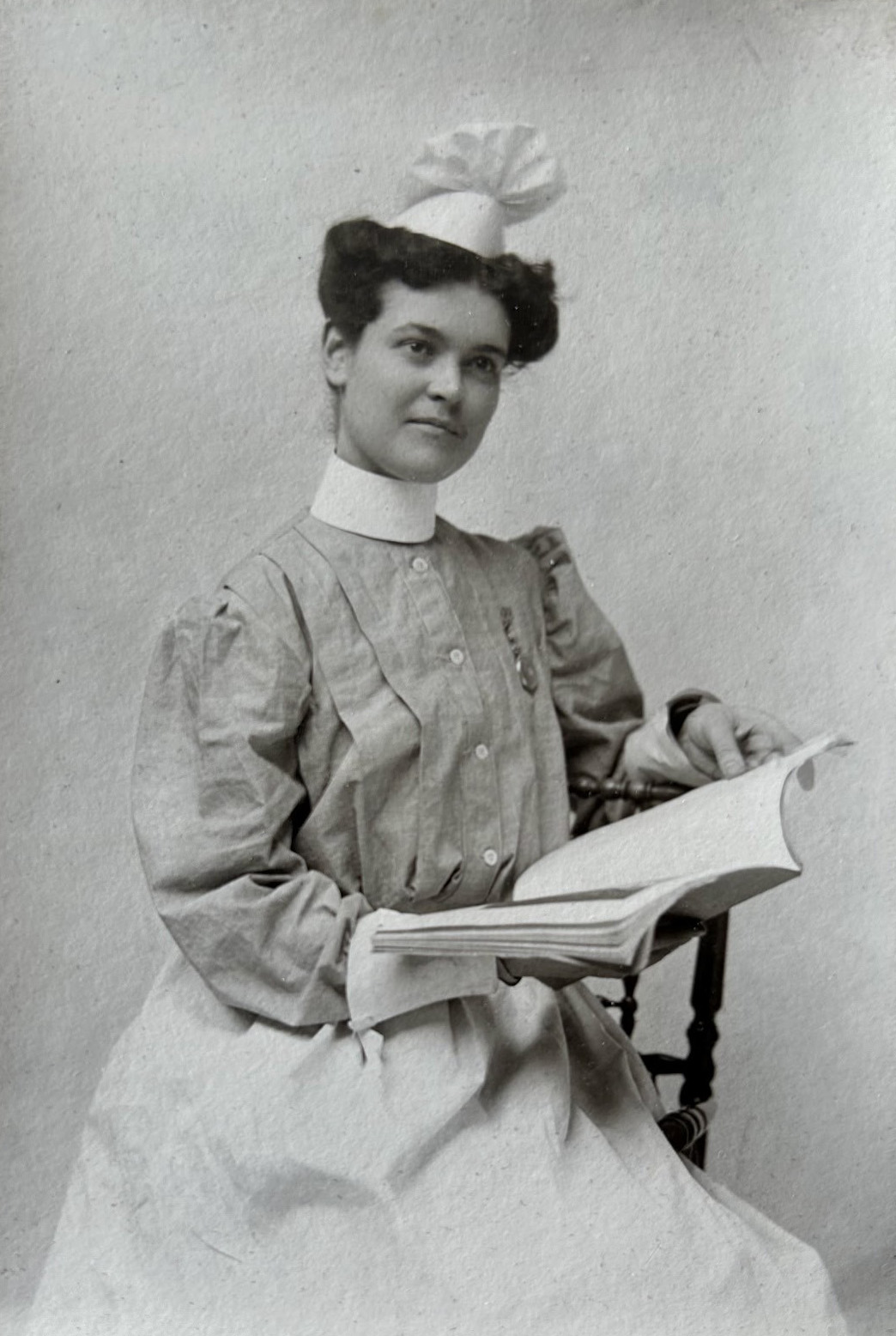 Black and white portrait photo of a white woman in an early 1900s nursing uniform and hat holding an open book