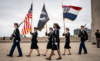 Modern photo of six young people in military uniform marching in a line carrying flags, rifles or swords