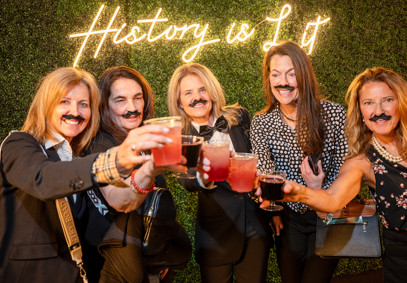 Modern photo of five white women wearing stick-on mustaches clinking their drinks together. Photo backdrop reads 'History is Lit'