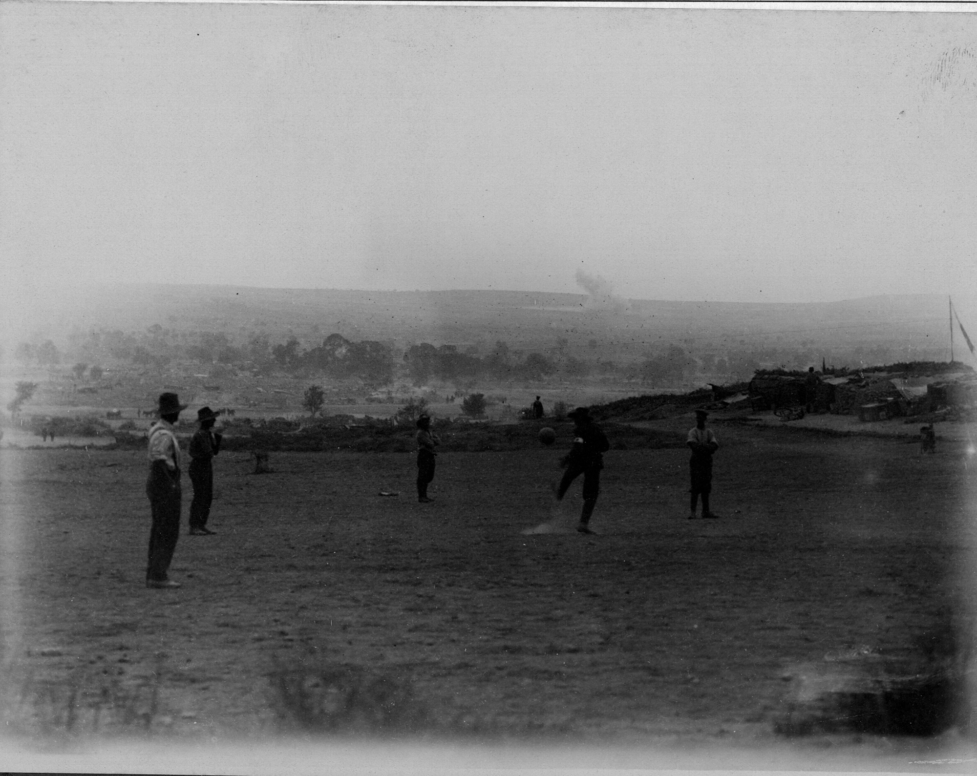 Black and white photo of a group of WWI soldiers playing football (soccer) in an open field as shellfire explosions go on in the distant background