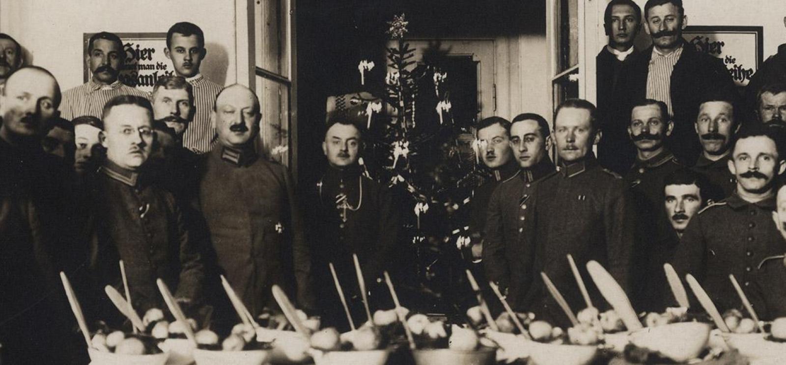 Large group of white men in military uniform gathered around an expansive table filled with food and utensils. There is a Christmas tree in the background.
