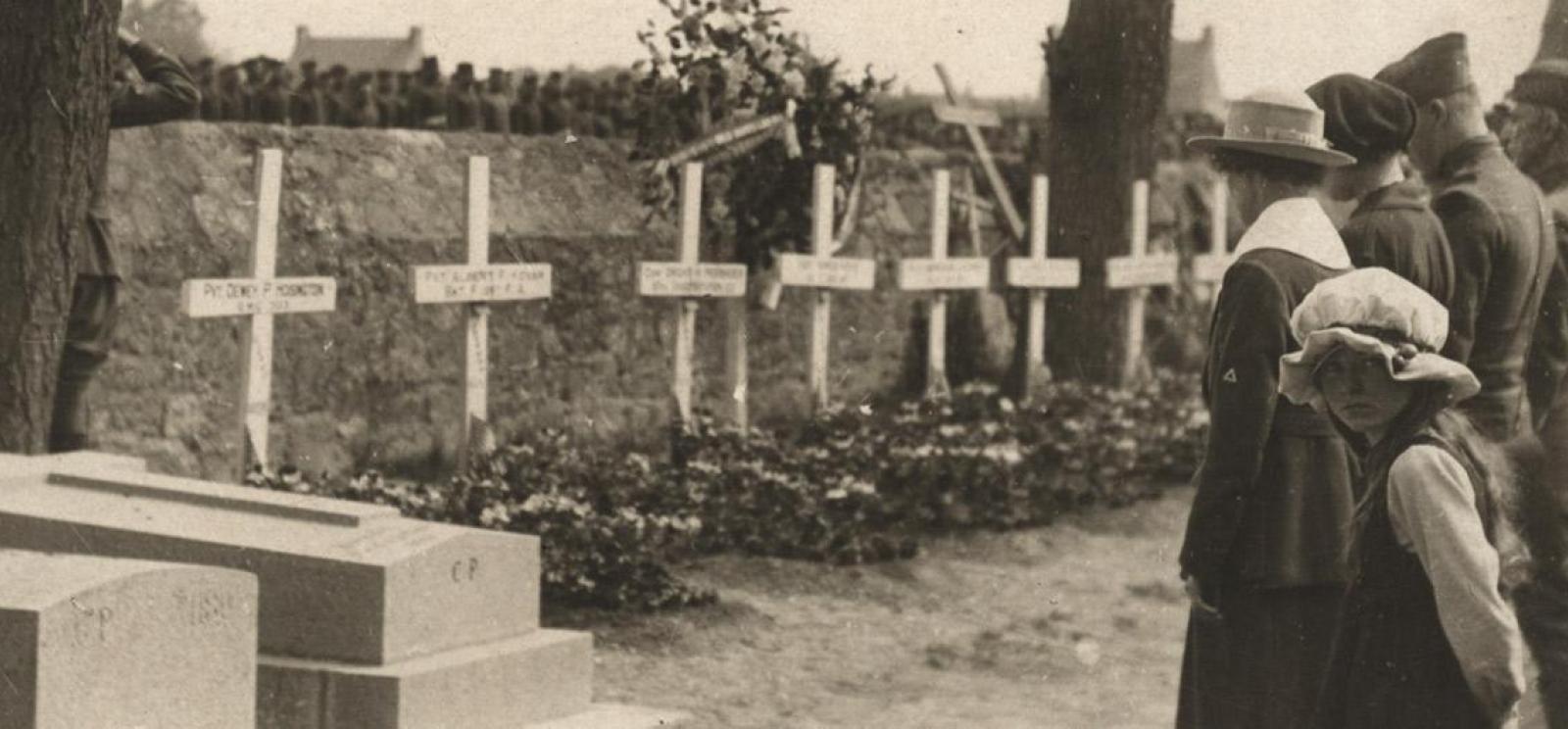 Sepia photograph of a row of people standing in front of a row of crosses. In the foreground a young girl in a large hat turns to look at the viewer.