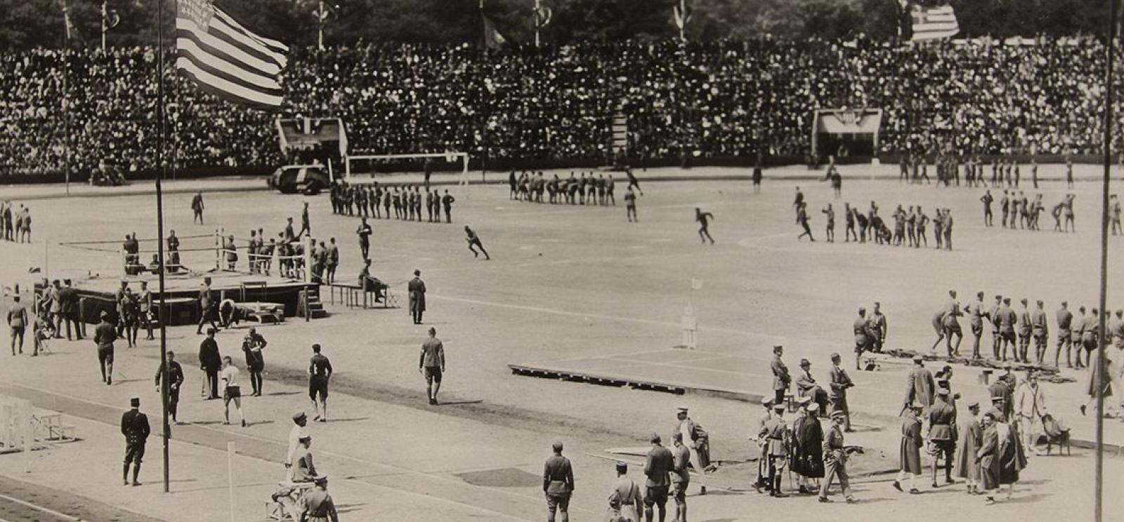Black and white photograph. View of a wide open sports field in a stadium filled with spectators. Different track and field events are taking place in different spots on the field. 