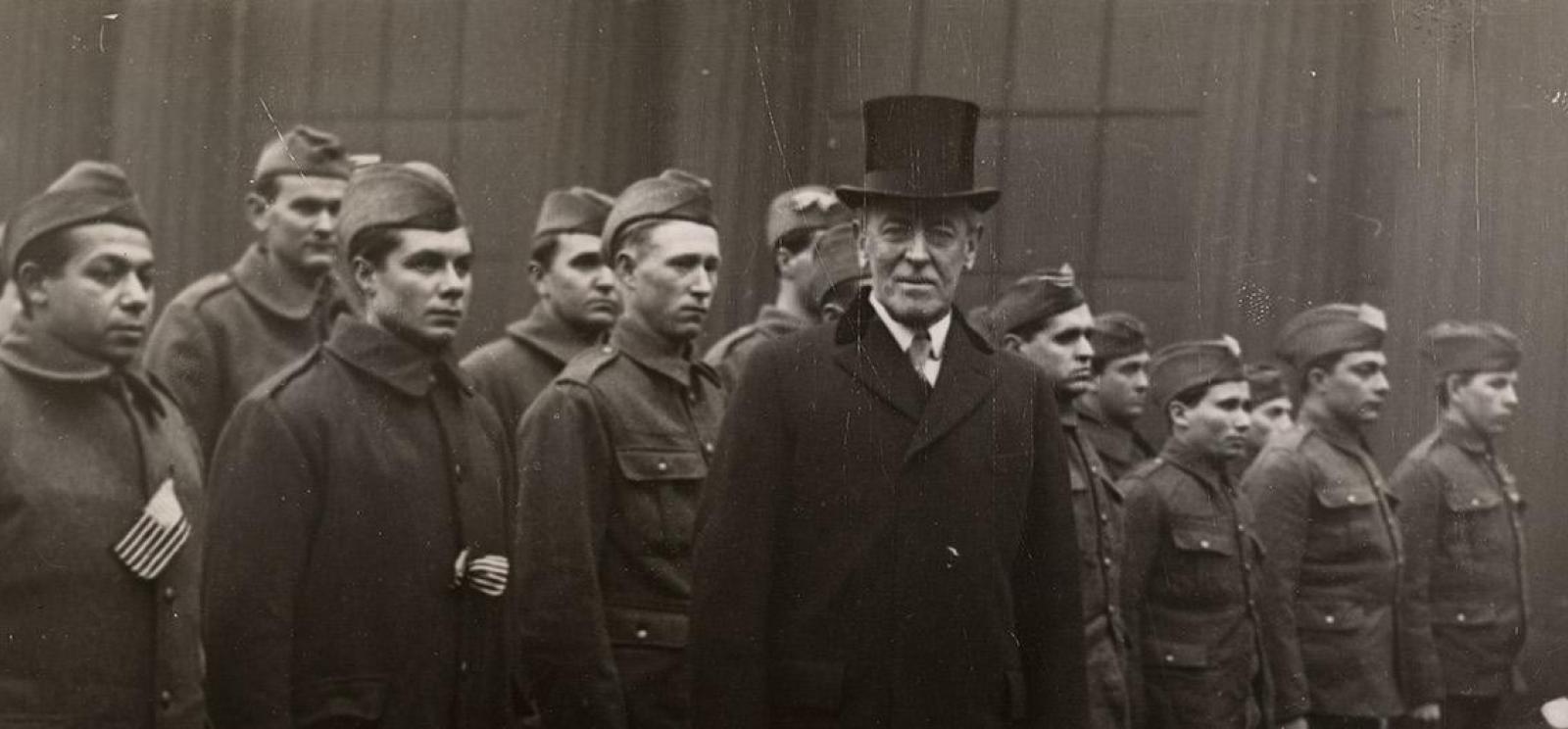 Black and white photo of Woodrow Wilson in a top hat standing in front of a line of soldiers in military uniform.