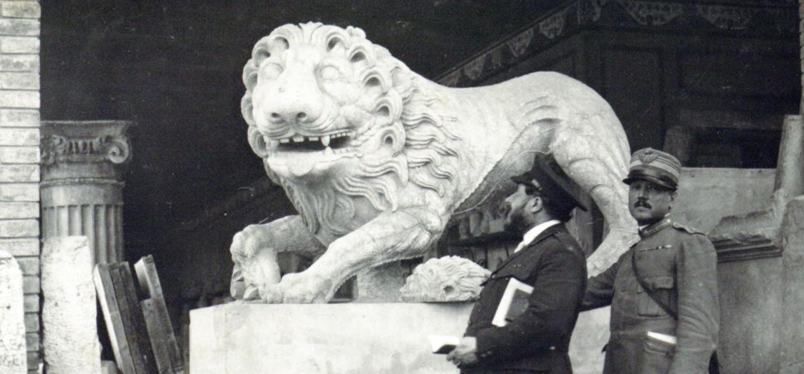 Photographie en noir et blanc d’un homme en costume debout devant une grande statue en pierre représentant un lion.