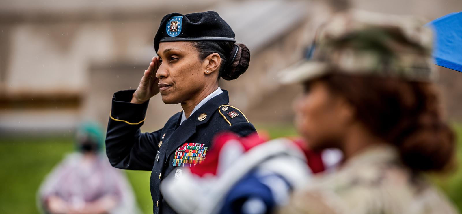 Modern photograph of a Black woman wearing military dress uniform saluting something out of frame. Another Black woman in camouflage uniform stands in the foreground out of focus.