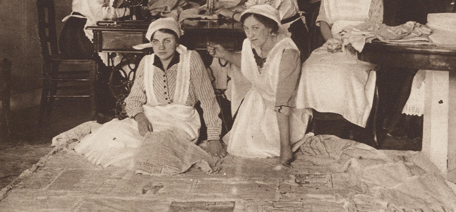 Black and white photograph of two white women in white pinafores kneeling on the ground surrounded by newspapers
