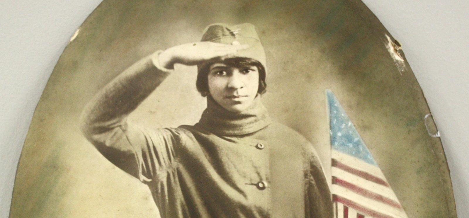 Sepia photograph of a Black woman in cap and uniform saluting while holding a U.S. flag.