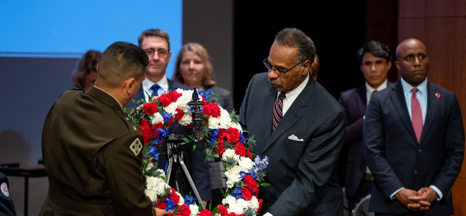 Modern photograph of a group of people wearing suits standing on a stage. In the foreground, an older Black man in a suit and a white man with a buzzcut wearing a military uniform place a large flower wreath on a stand.