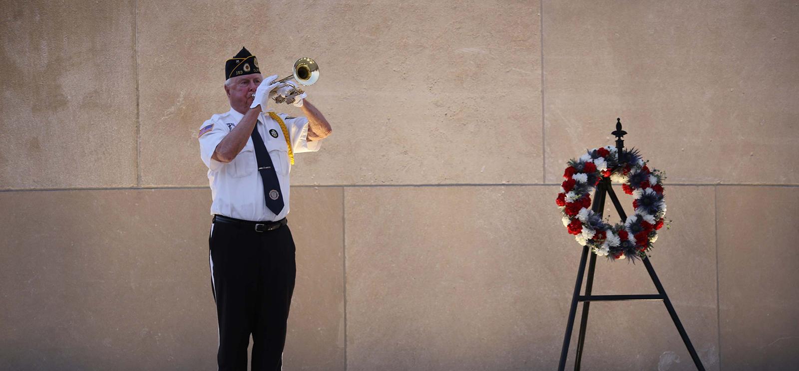 A uniformed bugler a playing Taps at the base of the Tower with a floral wreath in a stand to his right.