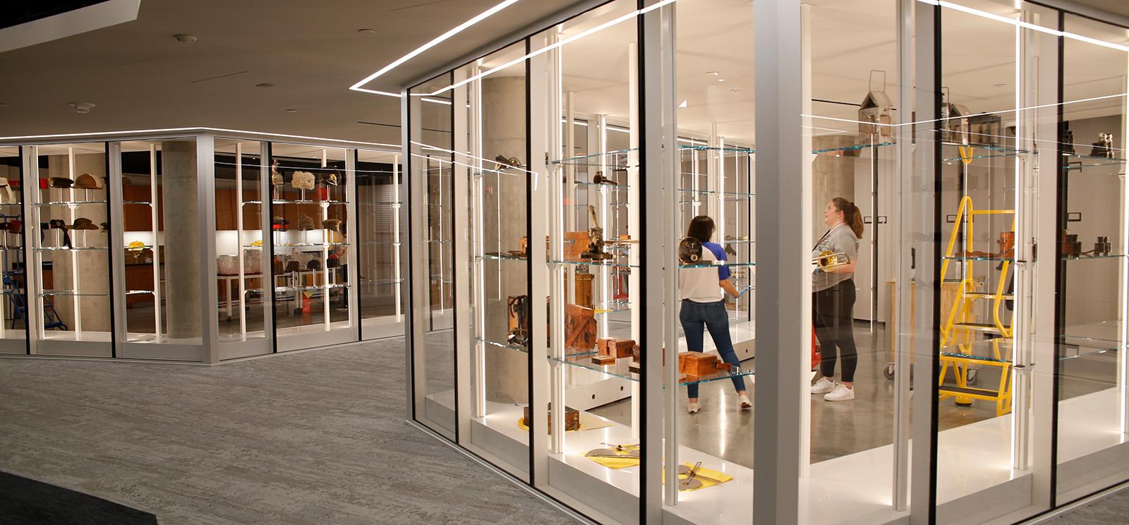 Modern photograph of a museum collections storage area as viewed through glass walls. Artifacts are also displayed along the glass walls. Two people stand in the storage area.