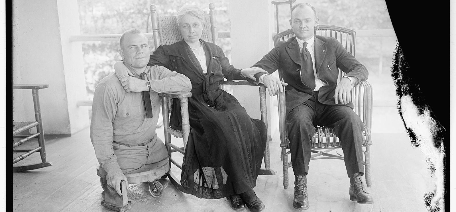 Black and white photo of three people posed for a group photo. Left to right: a white man in military uniform with both legs amputated nearly at the hip, resting on a small platform with wheels. A white woman in a dark-colored dress sitting in a rocking chair. A white man wearing a suit sitting in a rocking chair with a crutch leaning on a pillar behind him. The woman has her arm around the shoulders of the man on the left and her hand on the arm of the man on the right.