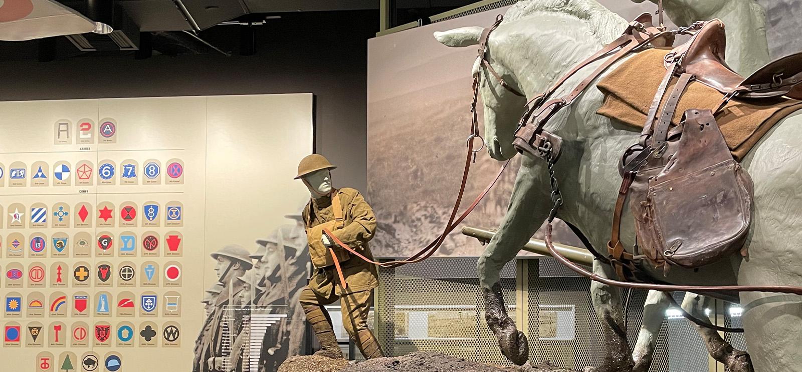 Modern photograph of a historical museum exhibit. A sculpture of a man dressed in WWI uniform struggles to lead several sculptures of mules through muddy terrain.