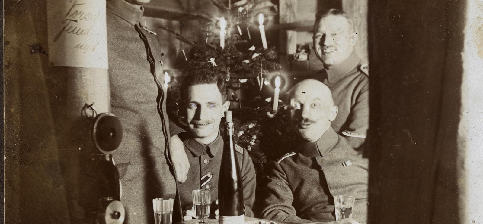 Black and white photograph of several WWI soldiers sitting in front of a Christmas tree with smiles on their faces and drinks on the table