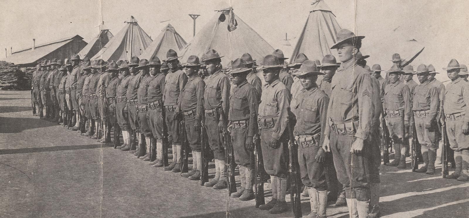 Photographie en noir et blanc de deux rangées de soldats amérindiens au garde-à-vous devant des tentes et des casernes.