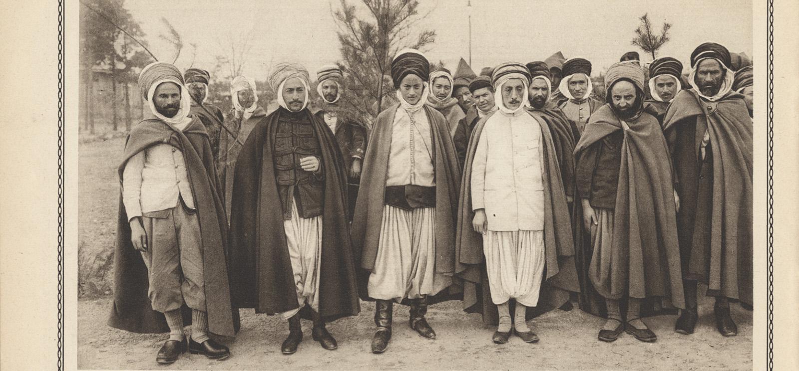 Crop of a scan of a vintage magazine page featuring a photograph of a large group of Muslim men in loose robes and head coverings, posed for a standing group portrait.