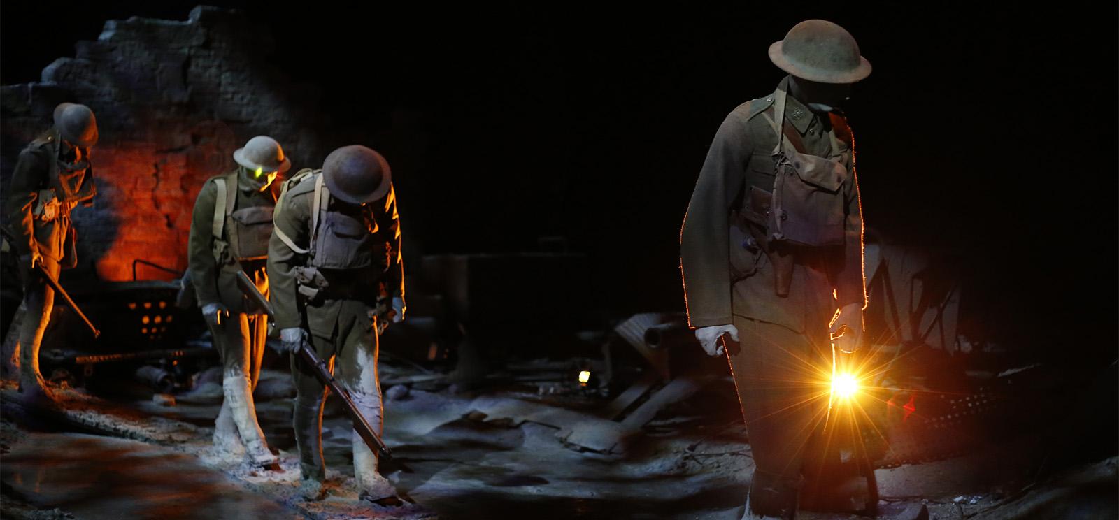 Photographie moderne de mannequins de soldats grandeur nature de la Première Guerre mondiale marchant péniblement sur un paysage de bataille boueux, éclairé de manière austère par des projecteurs orange et blancs