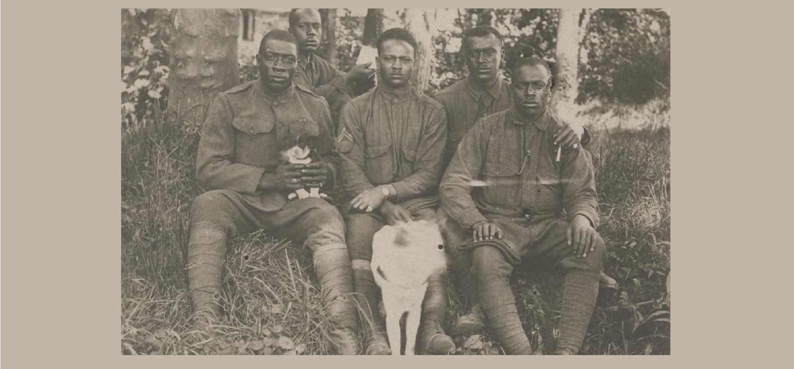 Black and white postcard photograph of five African American soldiers seated casually in wooded area with puppy on one soldier's lap and another dog in front center. 