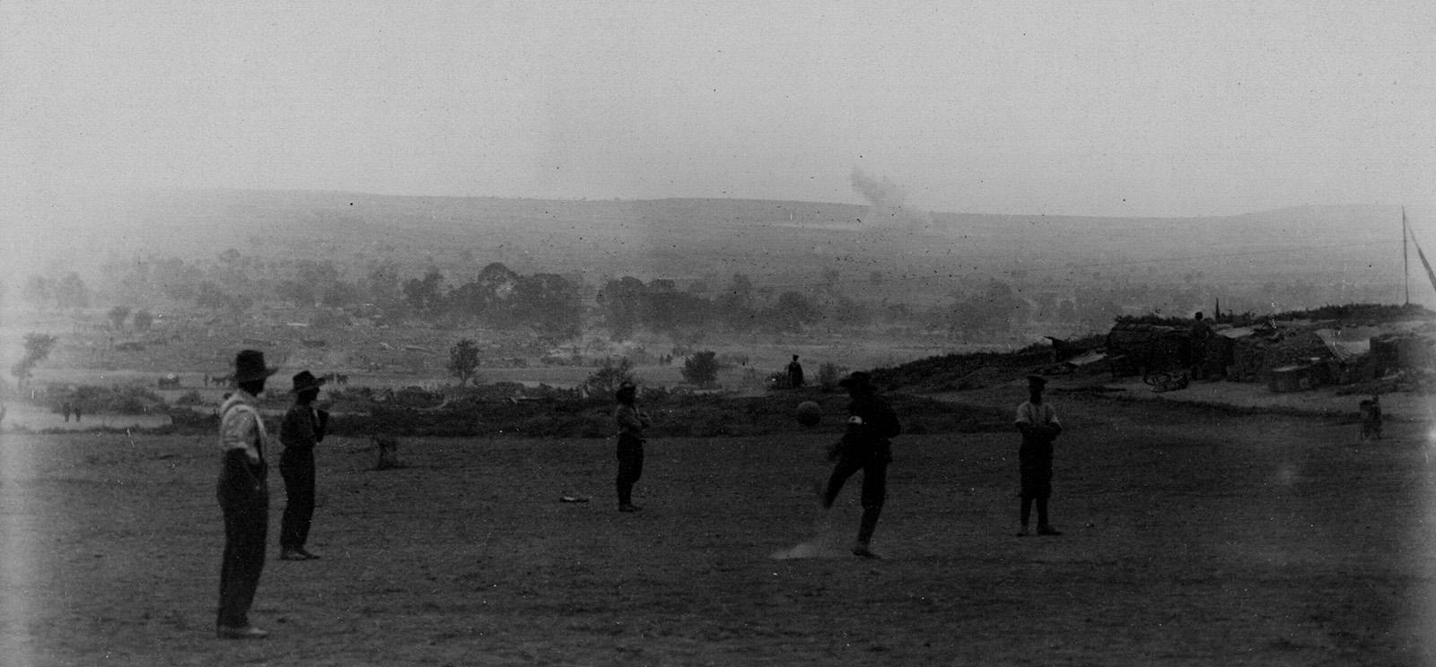 Black and white photo of a group of men in a field playing football (soccer). A bombshell explosion is going off in the distant background.