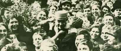 Black and white photograph of a happy crowd. In the center, a woman kisses the cheek of a man in a military cap.